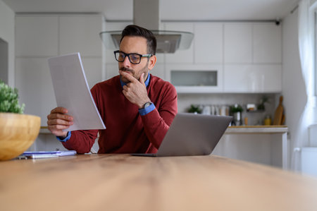 Focused male entrepreneur analyzing business report and working over laptop on desk in home officeの写真素材