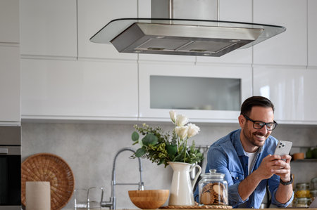 Smiling male entrepreneur online messaging over smart phone while leaning on kitchen counterの写真素材