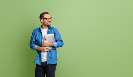 Young smiling businessman holding digital tablet and thinking while looking away on green backgroundの写真素材