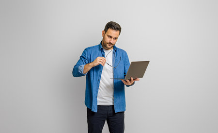 Thoughtful young businessman holding eyeglasses and looking at laptop isolated on white background