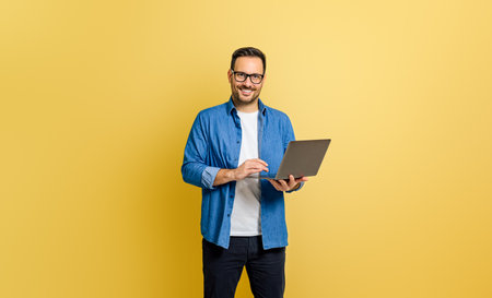 Handsome entrepreneur smiling and analyzing business report over laptop against yellow backgroundの写真素材