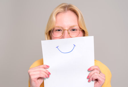 Portrait of young woman in eyeglasses holding white paper with smiley drawing on white background