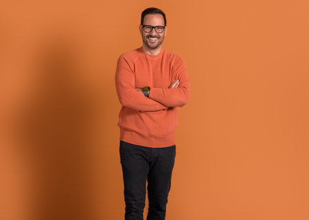 Successful young businessman in eyeglasses smiling and posing with arms crossed on orange backgroundの写真素材