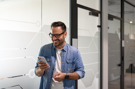 Happy male professional checking e-mails over mobile phone while standing by glass wall in office