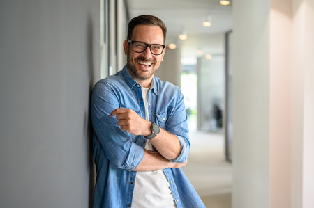 Portrait of positive male business leader in eyeglasses laughing and leaning on wall in modern officeの写真素材