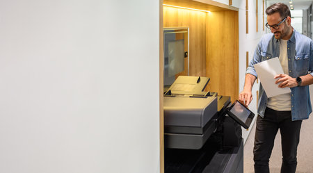 Male assistant in glasses holding financial documents and operating modern computer printer in...