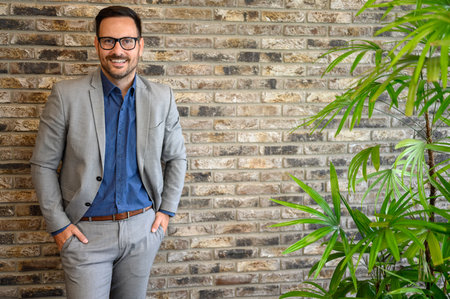 Portrait of confident sales manager with hands in pockets standing over brick wall in modern officeの写真素材