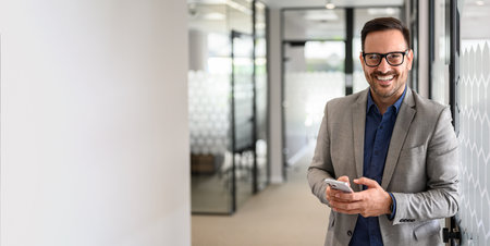Successful male CEO messaging over cellphone and looking at camera while standing in office corridorの写真素材