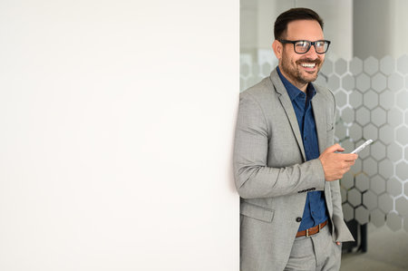 Portrait of smiling businessman using phone and looking away while standing by white wall in officeの写真素材
