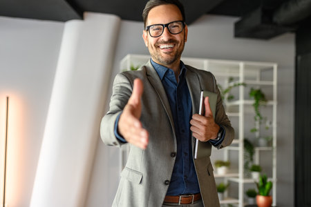 Portrait of confident happy businessman in eyeglasses holding laptop and offering handshake in officeの写真素材