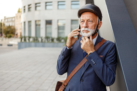 Portrait of serious old entrepreneur in helmet listening over phone call while standing by wall in cityの写真素材