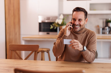 Smiling mid adult man with credit card talking over mobile phone while sitting at dining tableの写真素材