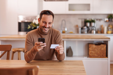 Portrait of mid adult man smiling and shopping online with credit card over smart phone at dining tableの写真素材