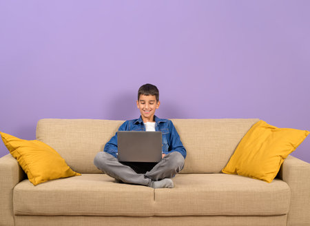 Smiling young schoolboy analyzing data over laptop while sitting on sofa against purple backgroundの写真素材