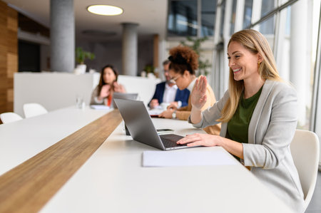 Smiling businesswoman waving at laptop during video call while colleagues working in the background...