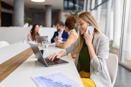 Smiling female entrepreneur working on laptop and discussing project details over call in meeting with professionals collaborating in the backgroundの写真素材