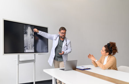 Male doctor discussing x-ray over computer and explaining problems to female patient in medical...