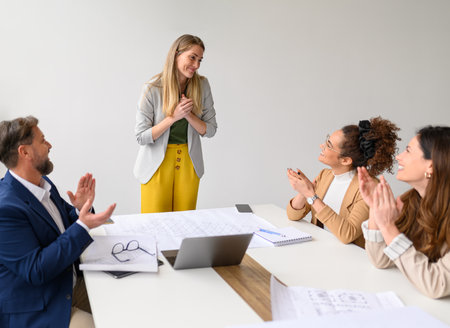 Happy businesswoman with hands clasped thanking professional colleagues applauding for her in office during meetingの写真素材