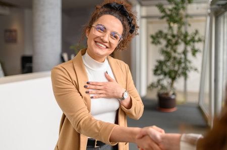 Smiling businesswoman in eyeglasses showing gratitude and shaking hands with female coworker in officeの写真素材