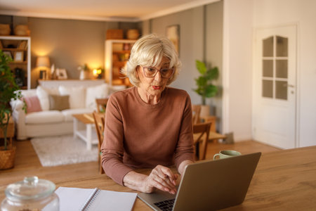 Senior woman in eyeglasses browsing products and shopping online over laptop while sitting at wooden...