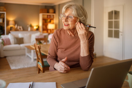 Elderly woman with glasses listening to doctor consultation on smartphone speaker while sitting at desk at homeの写真素材