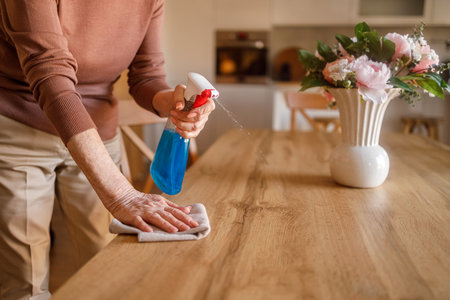 Midsection of elderly woman spraying cleaner on wooden dining table and wiping with napkin at homeの写真素材