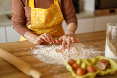 Midsection of elderly woman in apron mixing egg in flour while preparing content for food blog at...