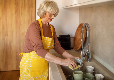 Elderly blond woman in eyeglasses and apron washing bowl in kitchen sink while enjoying household choresの写真素材