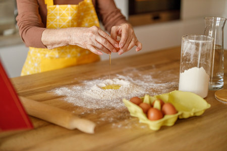 Midsection of senior woman cracking eggs into flour on kitchen island while blogging cookie recipes at homeの写真素材