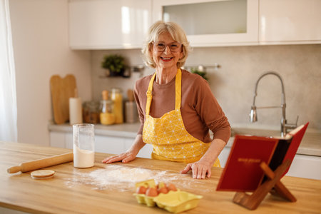 Smiling senior woman wearing apron with baking ingredients and recipe book placed on wooden kitchen island at homeの写真素材