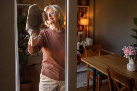 Elderly woman smiling and wiping glass door using napkin while doing household cleaning work at homeの写真素材