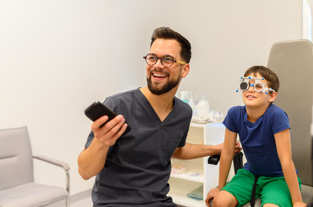Male eye specialist holding remote control testing eyesight of boy wearing phoropter during consultation at clinicの写真素材