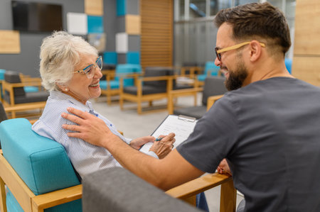 Male doctor comforting senior woman holding medical record while sitting together in hospital during visitの写真素材