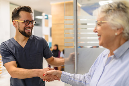 Male ophthalmologist opening glass door and greeting smiling senior woman with handshake at hospital entranceの写真素材