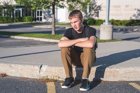 Sad teenage boy sitting on the curb in front of his school.の写真素材