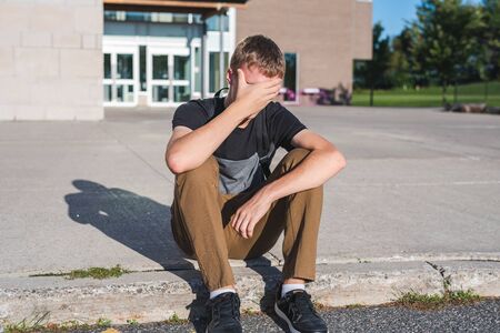 Sad teenage boy sitting on a curb while he covers his face with his hand.の写真素材
