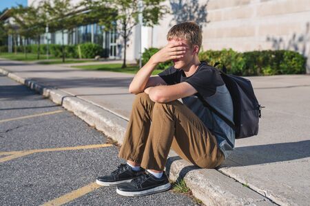 Sad teenage boy sitting on a curb while he covers his face with his hand.の写真素材