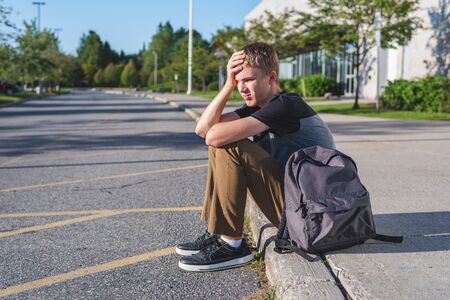 Upset teenager resting his head on his hand as he sits on the curb in front of his school.の写真素材