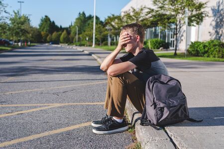 Sad teenage boy sitting on a curb while he covers his face with his hand.の写真素材
