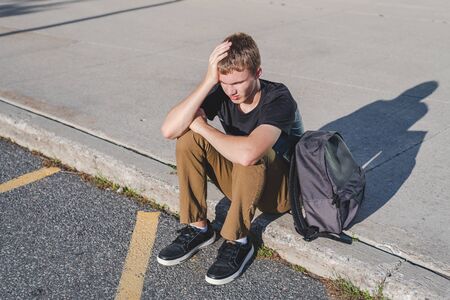 Upset teenager resting his head on his hand as he sits on the curb in front of his school.の写真素材