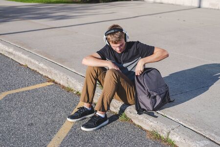 Happy teenager sitting on the ground with his backpack while listening to some music.の写真素材