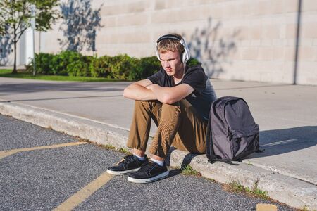 Sad teenage boy sitting on curb near school while listening to music on his headphones.の写真素材