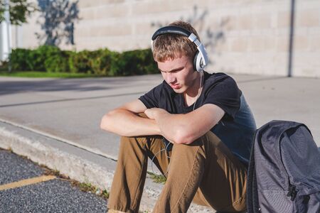 Sad teenage boy sitting on curb near school while listening to music on his headphones.の写真素材