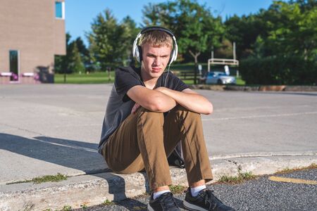 Sad teenage boy sitting on curb near school while listening to music on his headphones.の写真素材