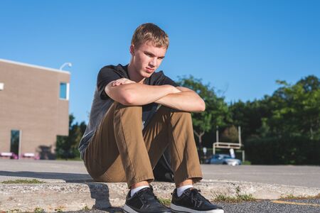 Sad teenage boy sitting on the curb in front of his school.