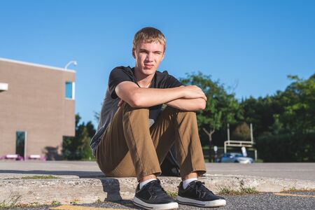 Sad teenage boy sitting on the curb in front of his school.の写真素材