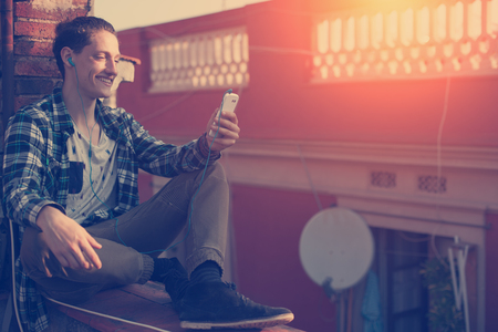 Young and smiling man sitting on the roof with mobile phone and listening musicの写真素材