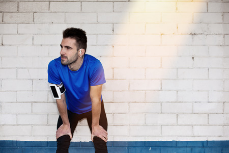 tired male athlete with earphones resting near brick white wall with sun lightの写真素材