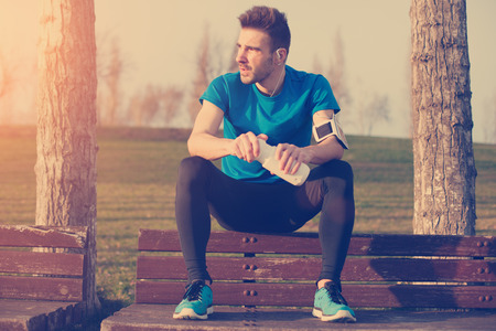 Handsome male athlete resting on the bench in park with bottle of water, armband with mobile phone and listening music (intentional sun glare and vintage color)の写真素材