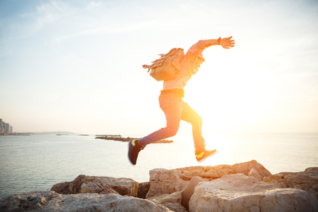brave man with backpack running over rocks near ocean in sunsetの写真素材